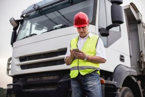 A truck driver in a red hard hat and high-visibility vest using a smartphone in front of a truck, highlighting the use of technology to prevent cargo theft.