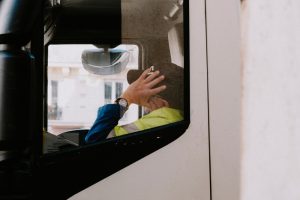 A truck driver sitting in the cab of a truck, seen through the window, wearing a high-visibility jacket and a cap, with one hand adjusting the cap. The image focuses on the challenges of truck driver retention, emphasizing the working conditions and environment inside the truck.