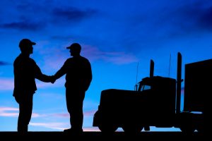 Silhouette of two truck drivers shaking hands with a semi-truck in the background during dusk. The image signifies collaboration and support among truck drivers, crucial for addressing truck driver health issues.