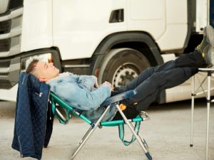 Young man resting on a folding chair in front of a truck, representing the journey to a truck driving job with no experience.