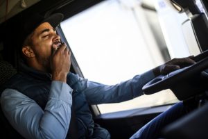 A truck driver wearing a cap and jacket yawns while driving, illustrating the signs of fatigue during long hauls.